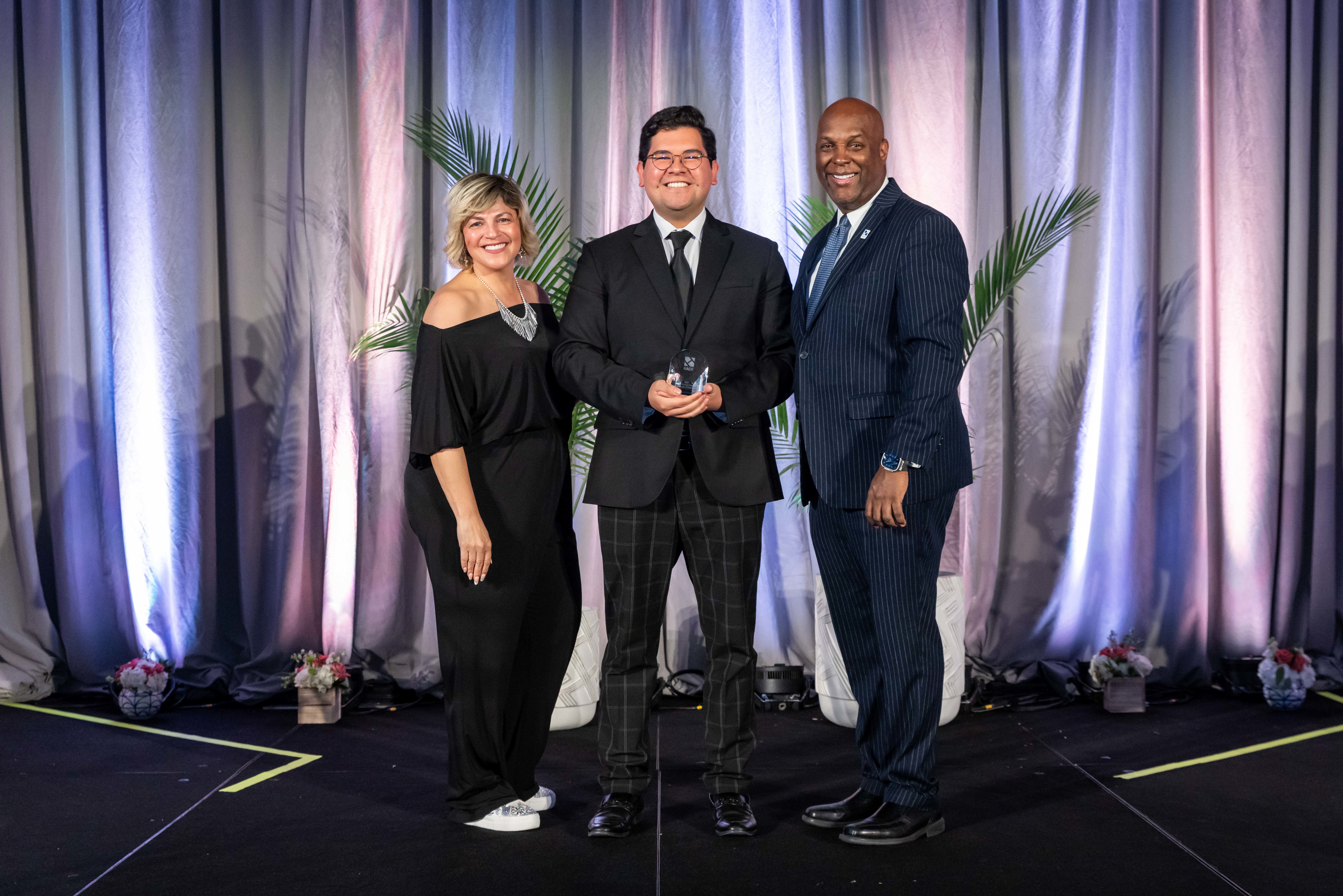 Man smiling with award, flanked by woman smiling in black jumpsuit and man smiling in blue pinstripe suit. 
