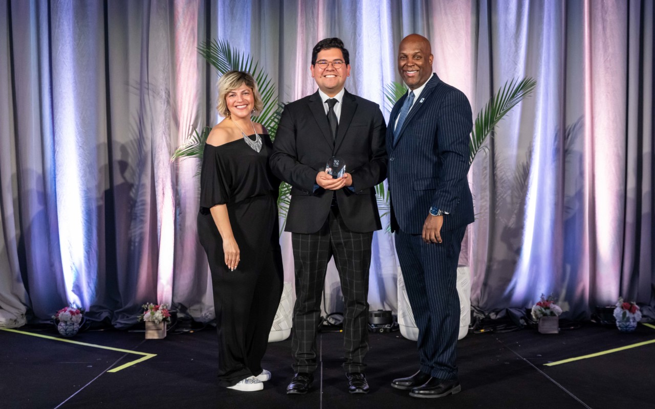 Man smiling with award, flanked by woman smiling in black jumpsuit and man smiling in blue pinstripe suit. 