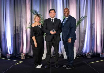 Man smiling with award, flanked by woman smiling in black jumpsuit and man smiling in blue pinstripe suit. 